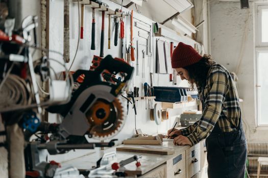 A man wearing a beanie focuses on a project in a tool-filled workshop.