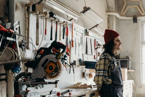 Free A man with long hair and beanie stands in a workshop with tools on the wall, reflecting a warm workspace atmosphere. Stock Photo