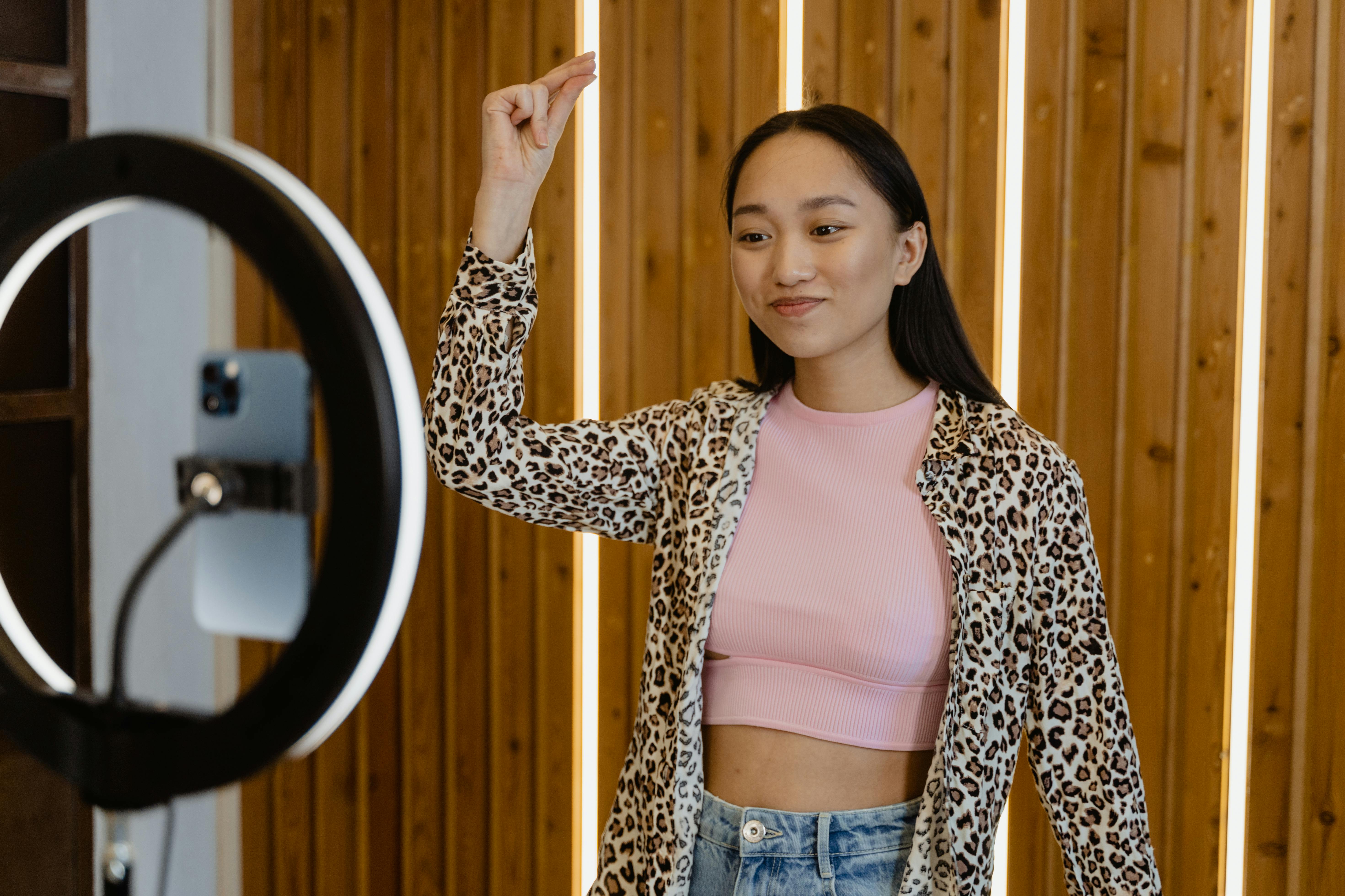 A smiling young woman records video using a smartphone and ring light indoors.