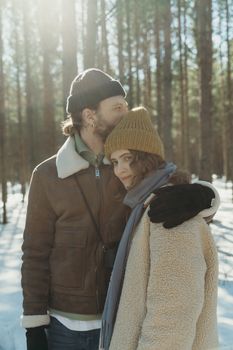 Couple embracing in a sunlit snowy forest wearing warm winter clothing, expressing love and togetherness.