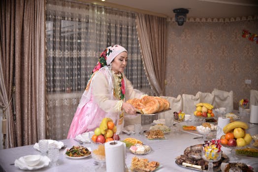 Woman in traditional attire sets a festive table indoors with various dishes, fruits, and bread.