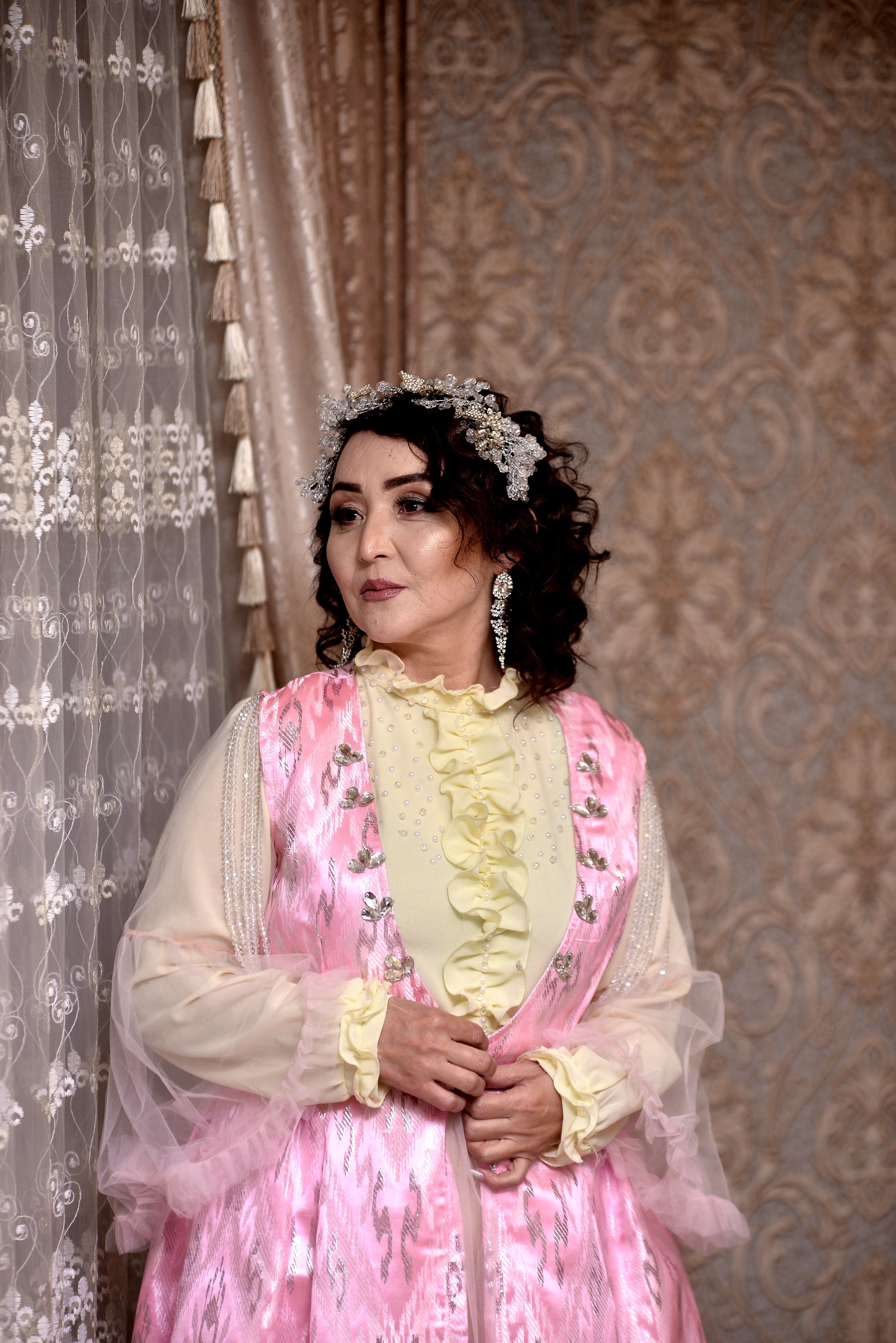 A woman stands near a window in elegant traditional attire with a pink dress and intricate headpiece.