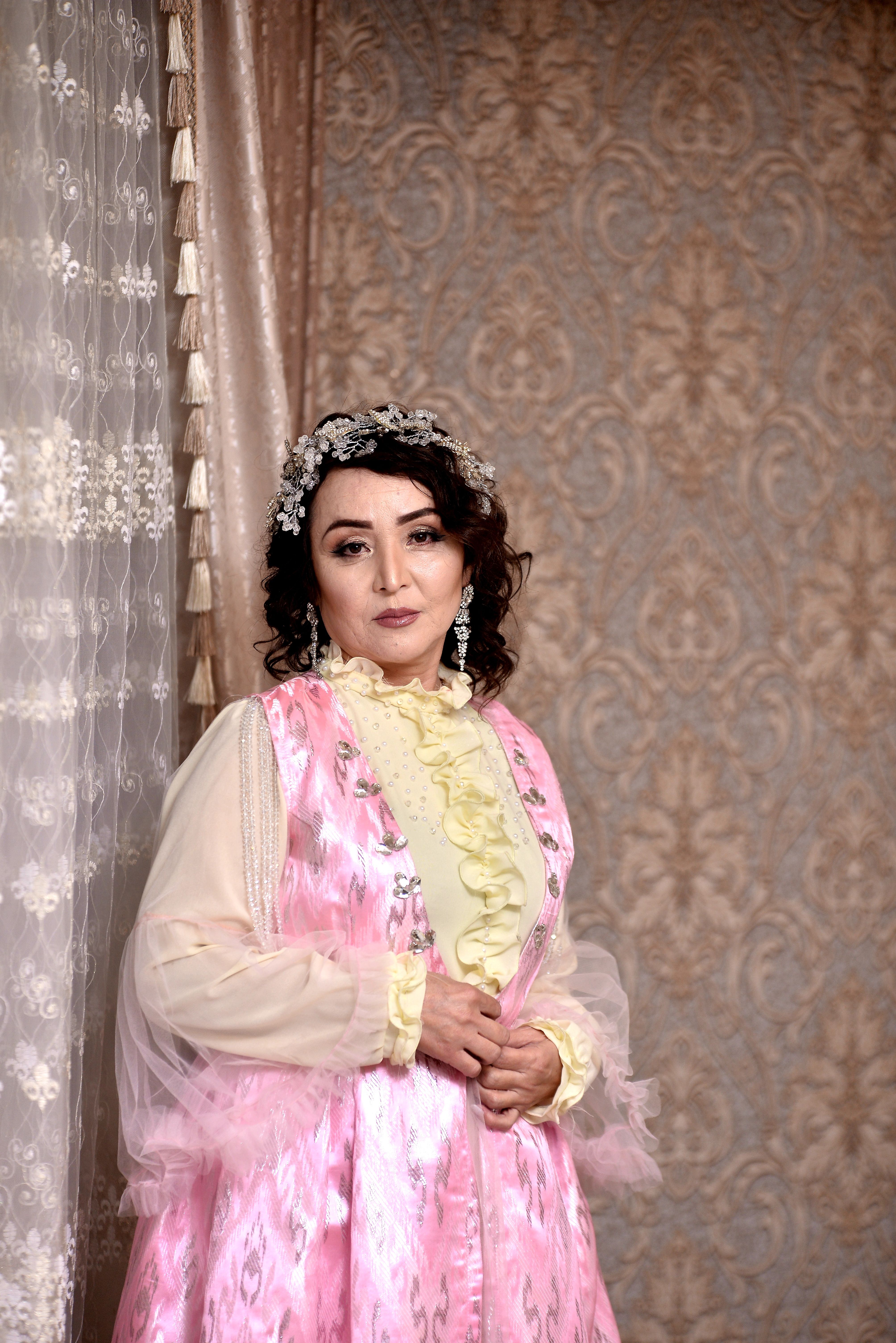 Portrait of an elegant woman in a pink traditional dress with floral headpiece, standing against ornate curtains.