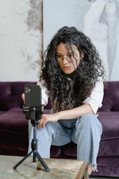 Woman with curly hair vlogging indoors, using smartphone on tripod, seated on couch.
