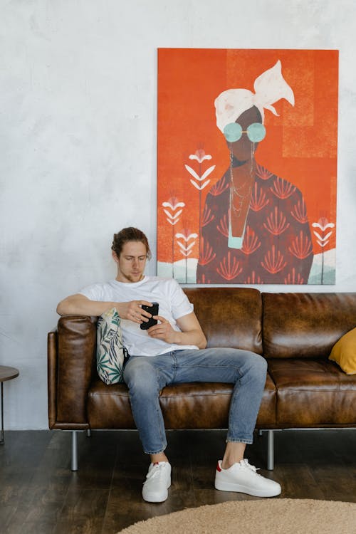 A Man in a White Shirt and Denim Pants Using His Smartphone while Sitting on a Couch