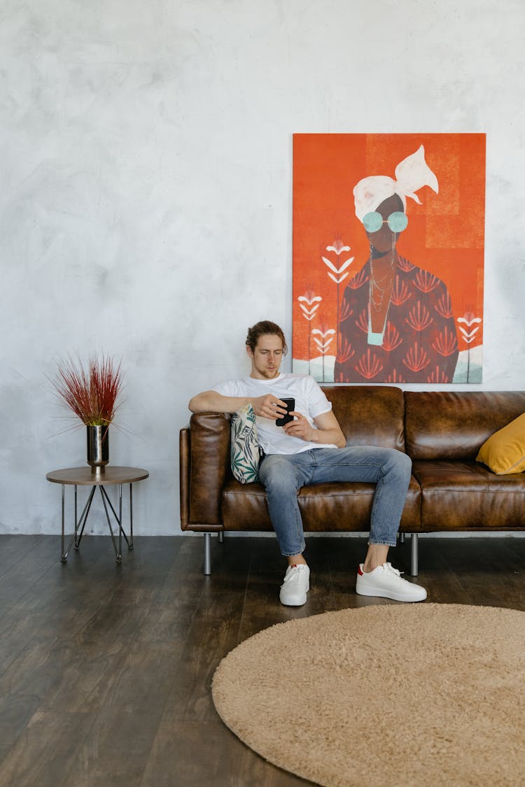 A Man In A White Shirt And Denim Pants Using His Smartphone While Sitting On A Couch