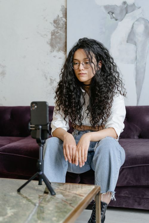 Free Woman with curly hair sits on a couch, vlogging with a smartphone indoors. Stock Photo