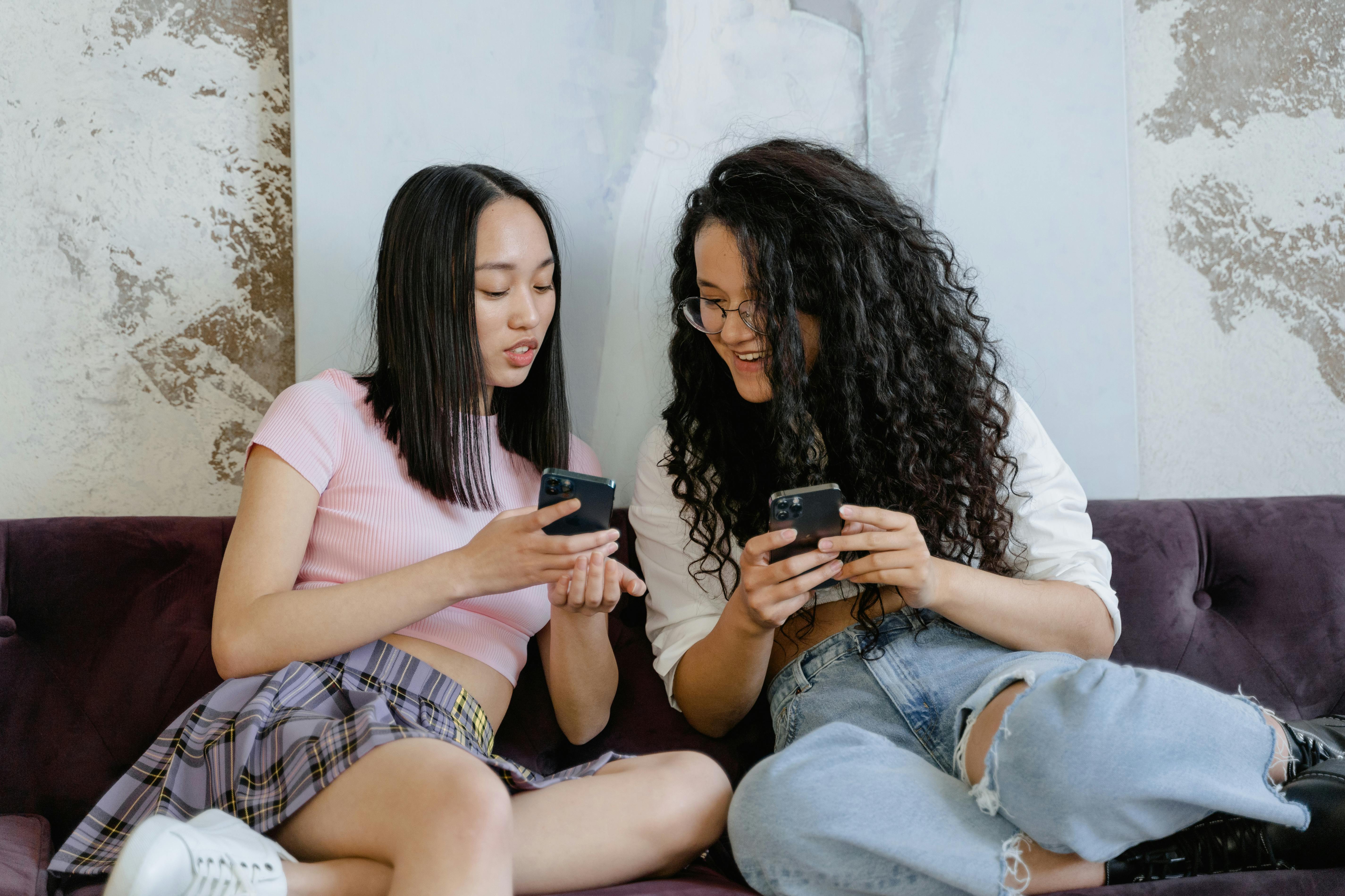 Two fashionable women sitting on a couch, engaged with their smartphones, sharing a moment.