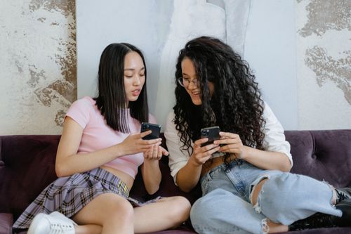 Free Two fashionable women sitting on a couch, engaged with their smartphones, sharing a moment. Stock Photo