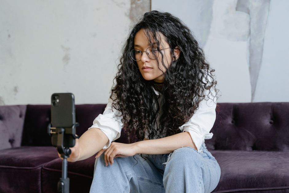 A young woman with curly hair sits on a sofa, using a smartphone mounted on a tripod.