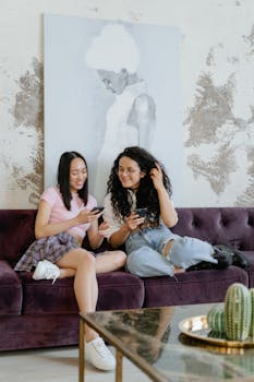 Two young women laughing and using smartphones on a cozy living room sofa.