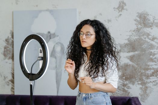 A young woman stands indoors, using a ring light and smartphone for streaming content creation.