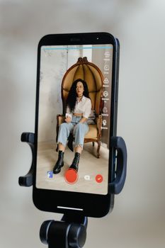 A woman with black hair sits on an ornate chair being streamed on a smartphone.