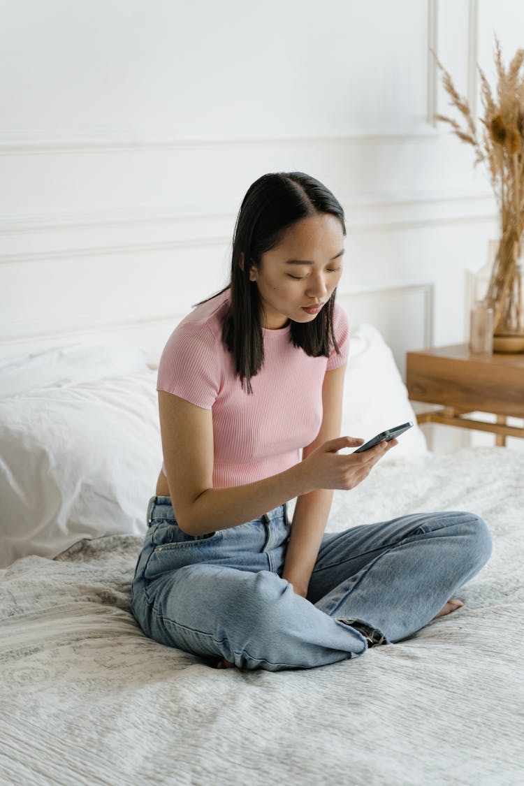 Woman In Pink Shirt Sitting On Bed While Holding A Cellphone