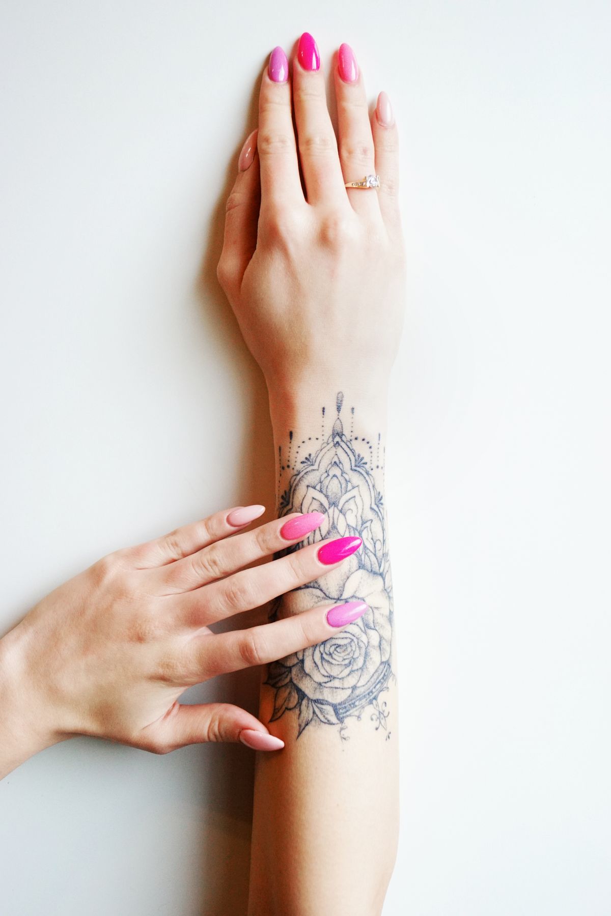 A close-up of tattooed arm and manicured hands with vibrant nail polish on a light background.