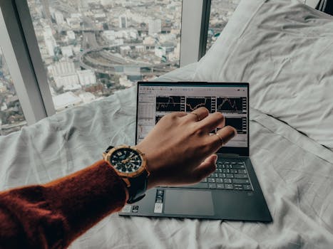 A person checks time while trading on a laptop with charts, overlooking a city view from bed.