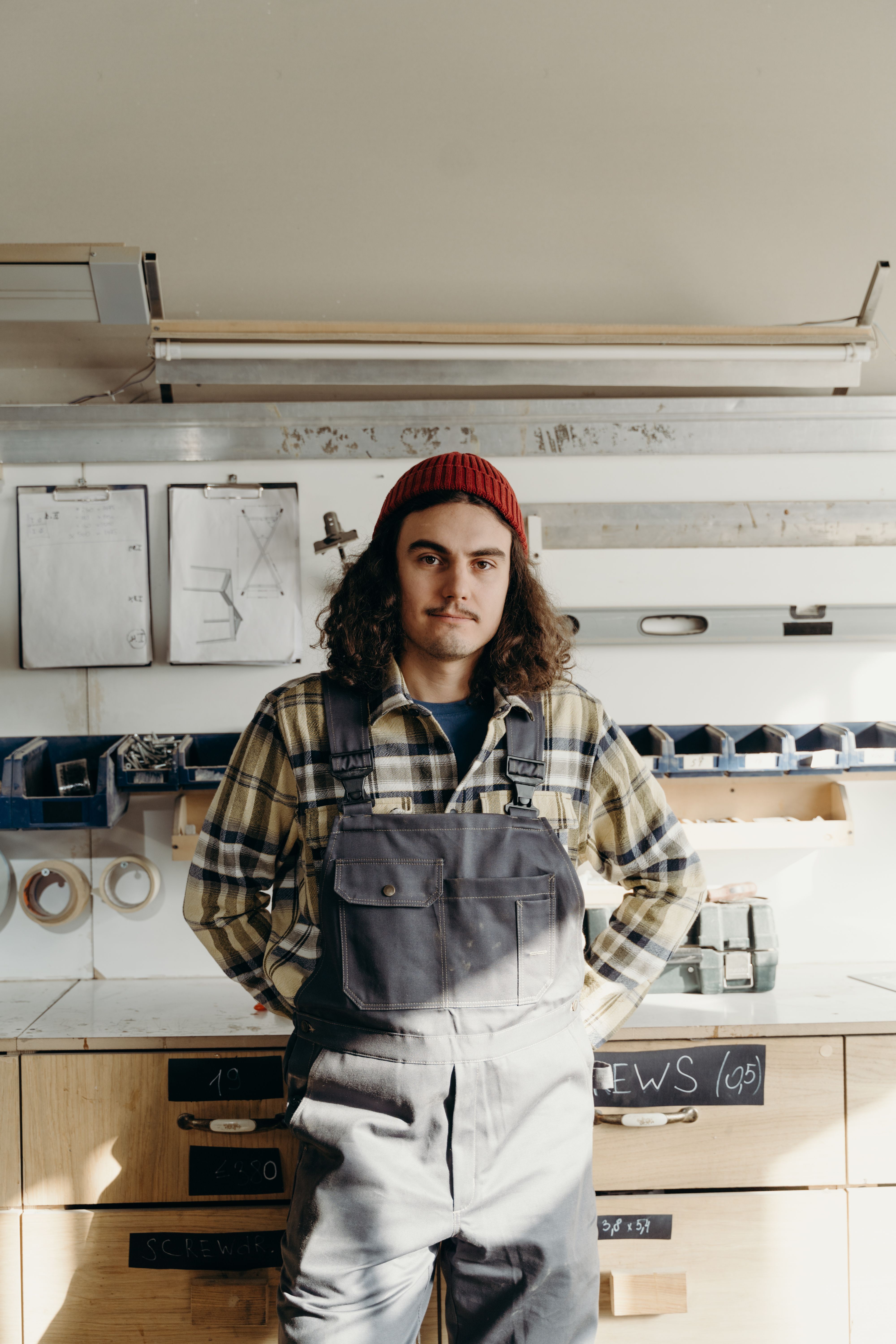 A craftsman stands confidently in a workshop, wearing a beanie and plaid shirt.