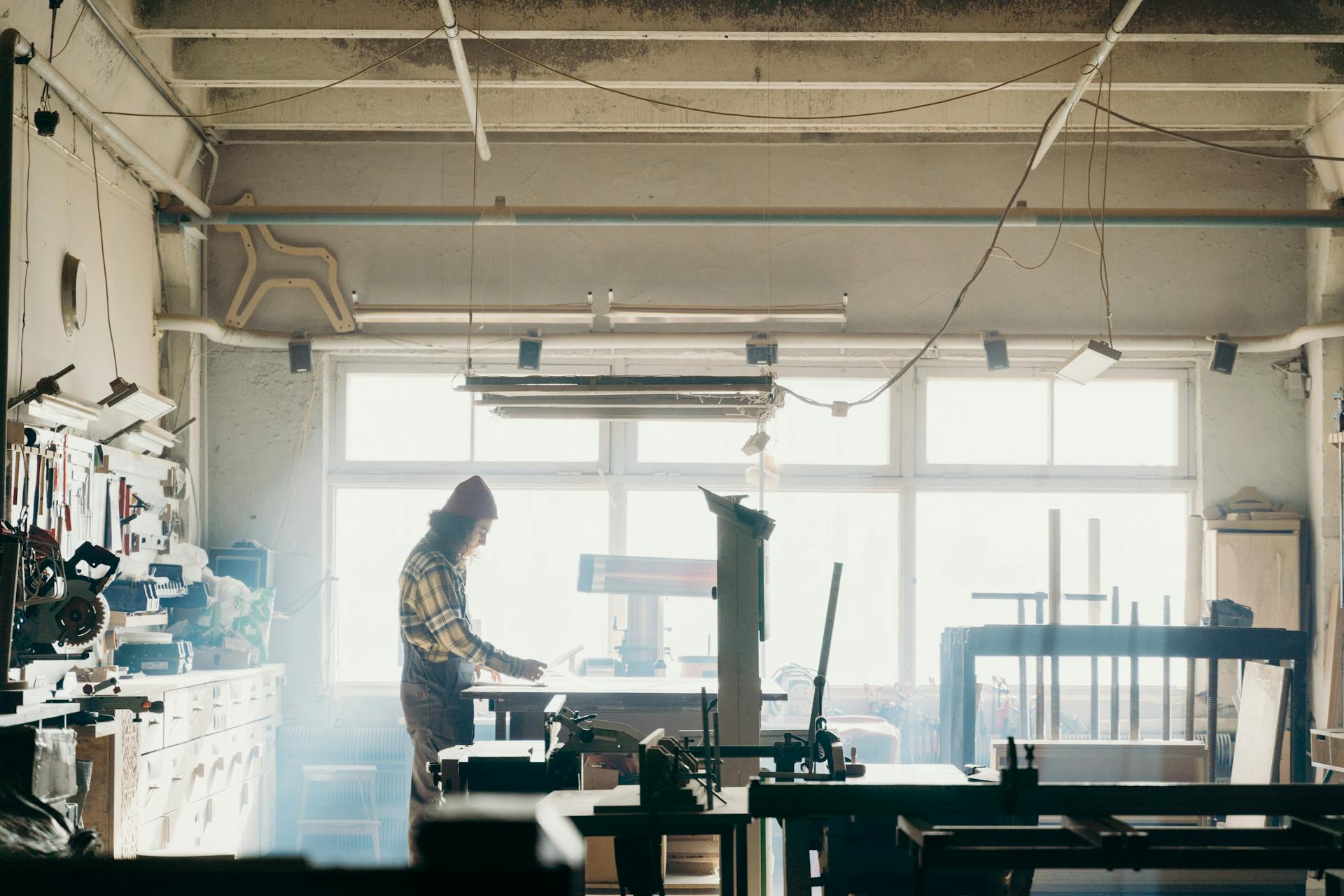 A craftsman focuses on woodworking in a sunlit workshop, surrounded by tools and machinery.