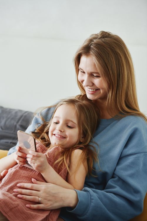 Free Caucasian mother and daughter smiling and using a smartphone at home, creating cherished family moments. Stock Photo