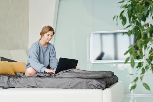 A woman in a sweatshirt works on a laptop while sitting on a bed in a contemporary bedroom setting.