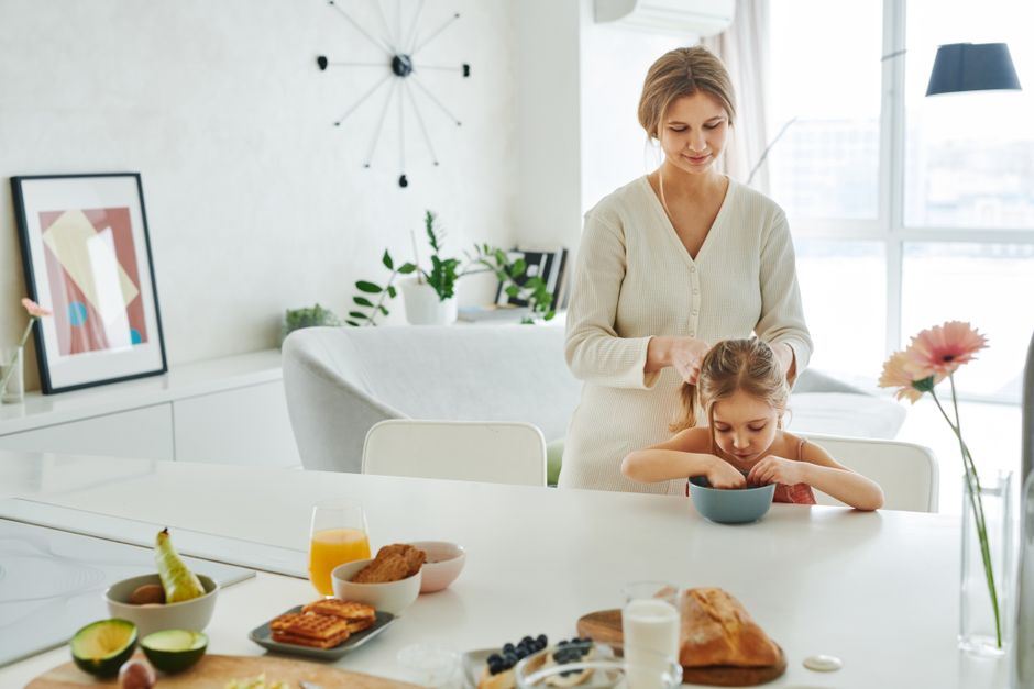 A mother braids her daughter's hair in a bright kitchen, surrounded by breakfast items. Uma mãe trança o cabelo da filha em uma cozinha bem iluminada, cercada por itens de café da manhã.
