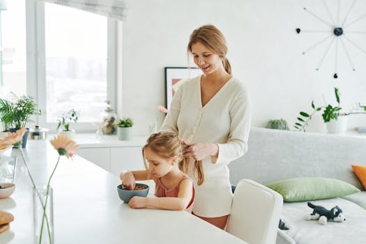A mother braiding her daughter's hair as the child eats breakfast at home in a cozy kitchen setting.