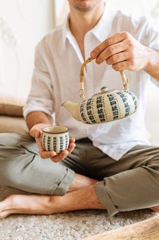 Adult man pouring tea from a decorative teapot while sitting cross-legged indoors.