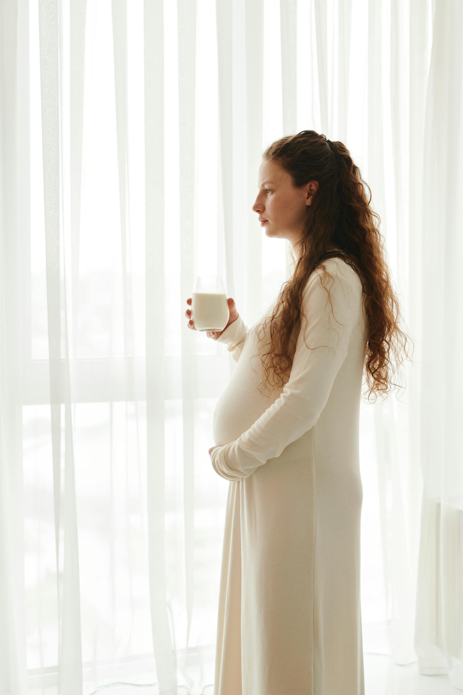 Expecting mother in white dress holding milk by a sheer window, highlighting maternal health.