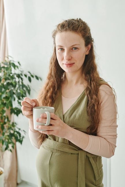 Mẹ Bầu Ăn Macca Úc Được Không? Nên Ăn Bao Nhiêu Là Tốt Nhất? 4 Pregnant woman smiling while holding a mug, standing in a bright indoor setting.