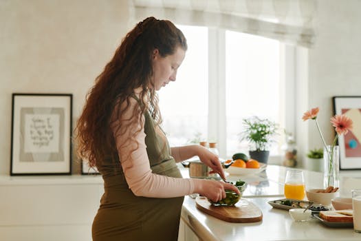 Pregnant woman with curly hair prepares a meal on kitchen counter, surrounded by fresh ingredients.