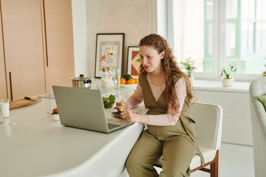 Pregnant woman using a laptop while sitting in a modern kitchen, enjoying a healthy snack.