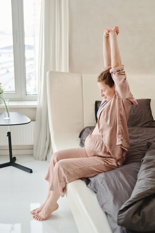 A Pregnant Woman in Pink Sleepwear Sitting on the Bed while Stretching Her Arms