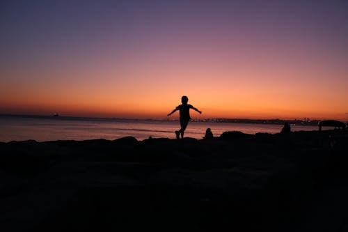 Back view of silhouette anonymous child running along sea shore against sunset sky with colorful gradient