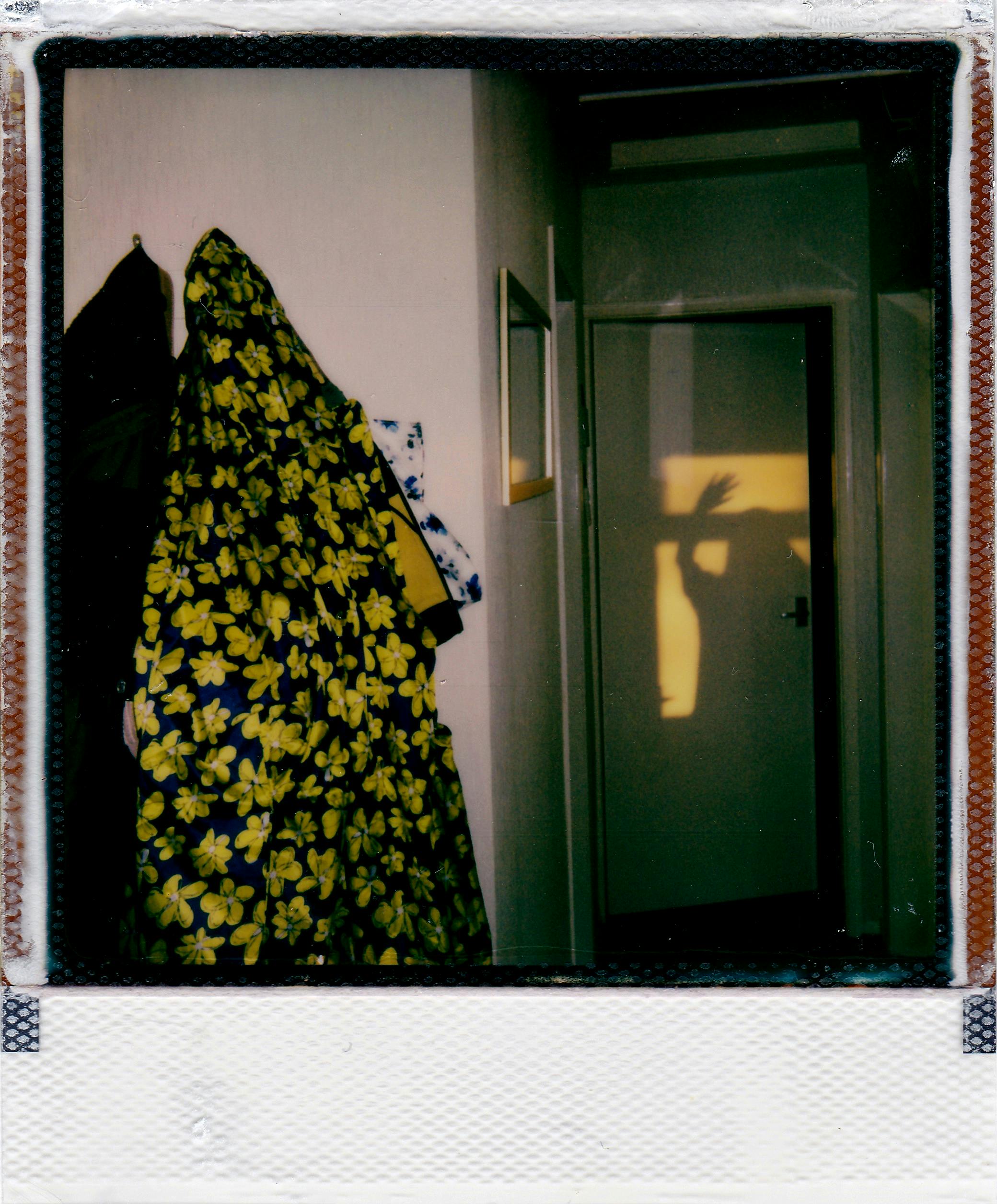 A vintage Polaroid image showing a shadowy figure in a hallway with colorful clothes hanging.