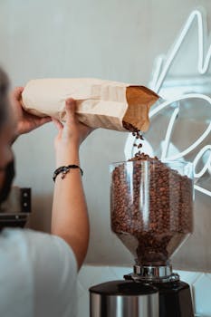 Barista pouring coffee beans into grinder from a paper bag inside a cafe.