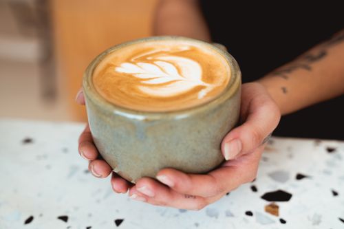 Free A close-up of a latte with leaf art in a ceramic mug, held by hands. Stock Photo