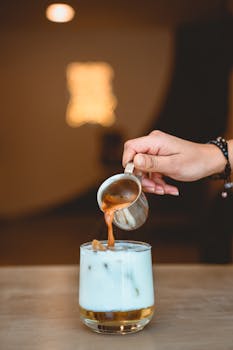 Hand pouring espresso over iced milk in a glass, perfect for a refreshing coffee drink.