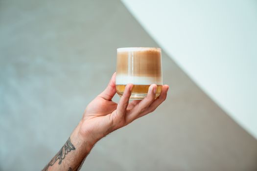 Hand holding a glass of layered coffee with a tattooed arm against a minimalist background.