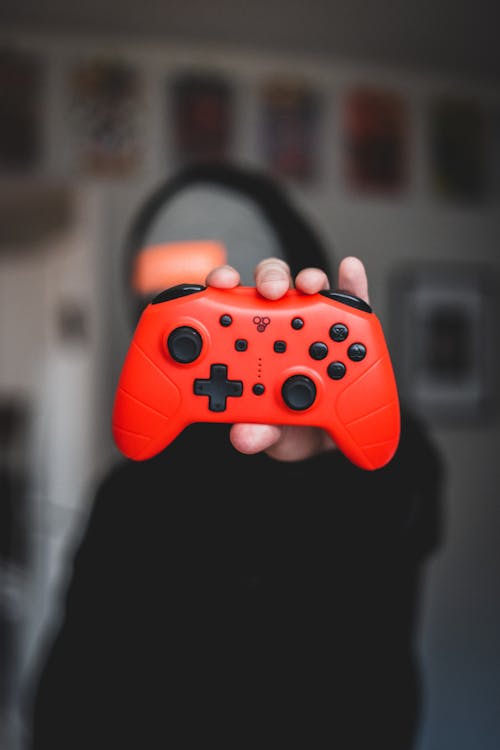 Blurred anonymous male gamer in black hoodie holding portable red gamepad with black buttons