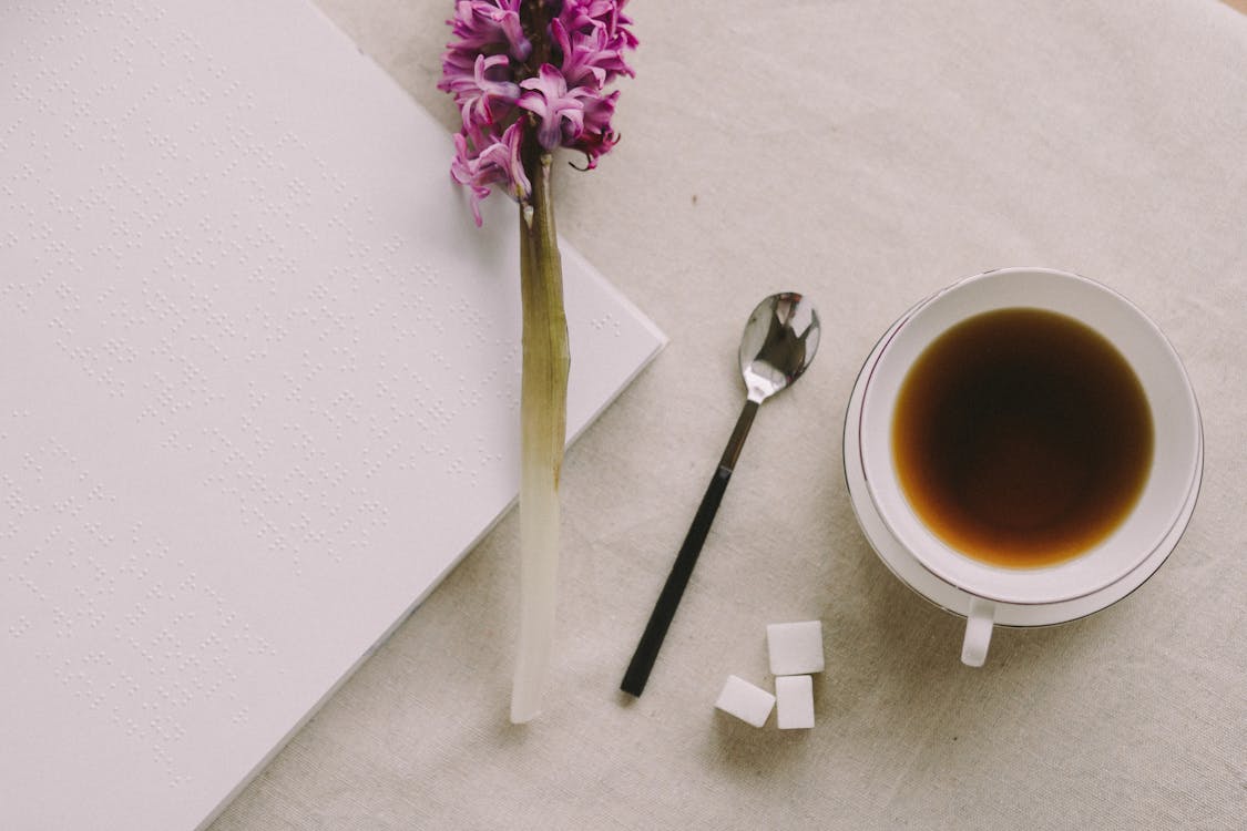 Free A minimalist flatlay featuring a coffee cup, braille book, flower, and spoon on a table. Stock Photo