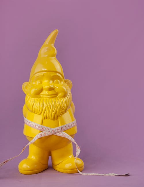 Composition of measuring tape on bright yellow garden gnome placed on left side on light purple surface