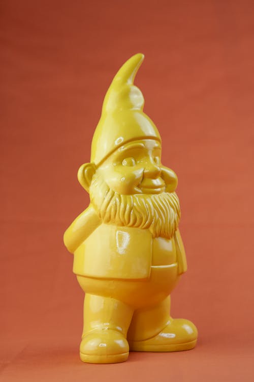 Small bright yellow ceramic garden gnome with beard and hat standing sideways on red background
