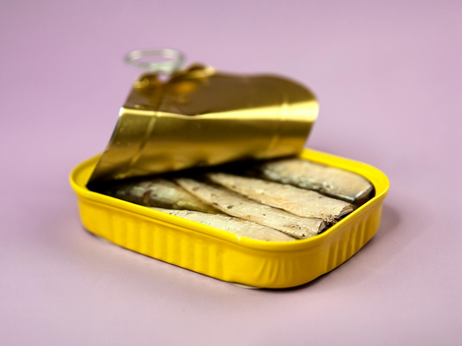 Opened delicious canned sardines in yellow container with oil on centre of light purple background