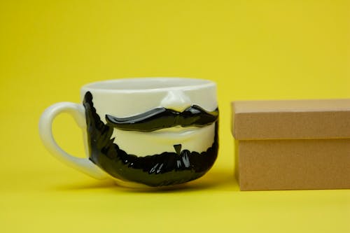 Porcelain cup with black beard and mustache near small cardboard box on bright yellow background