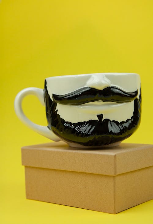 Composition of creative cup with mustache and beard placed on small cardboard box on yellow surface