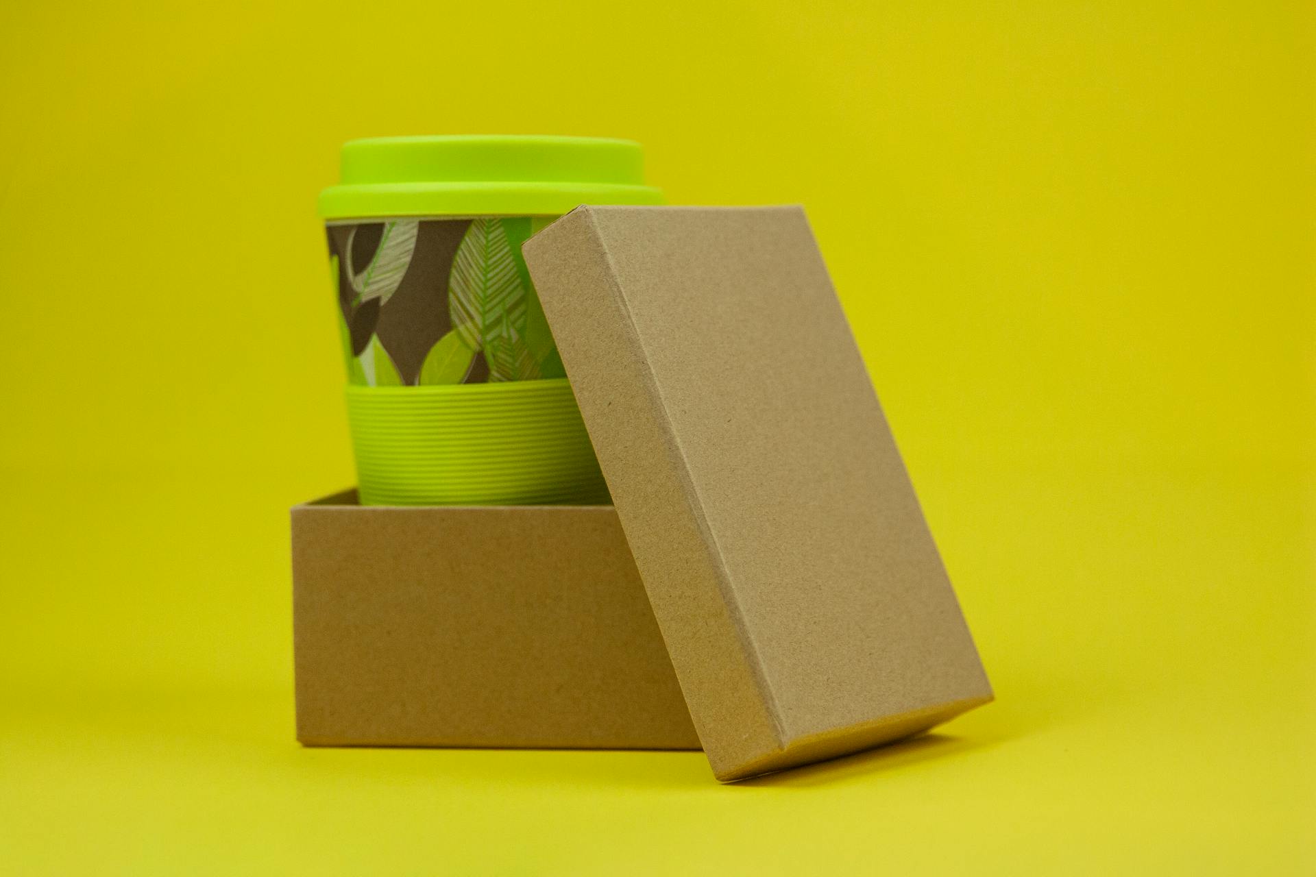 Reusable eco green bamboo cup with leaves in carton box on yellow background in light studio