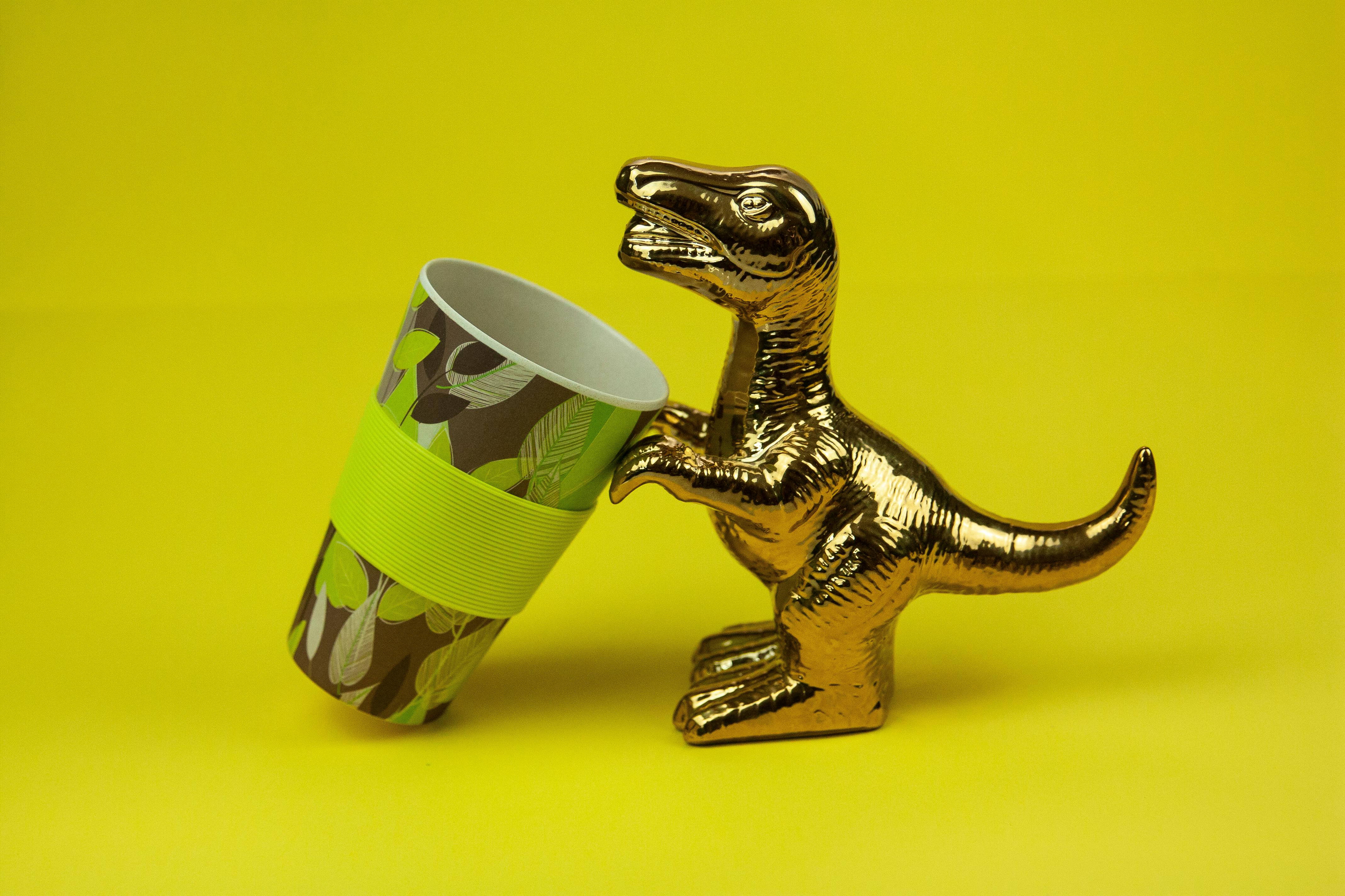 A golden dinosaur figurine holds a patterned cup against a vibrant yellow background.
