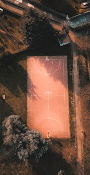 A drone captures a serene aerial view of a basketball court surrounded by trees at sunset, highlighting its natural beauty.
