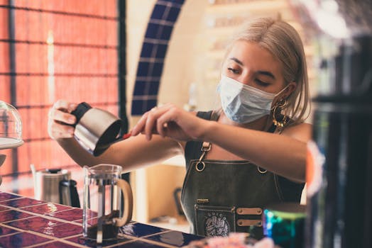A barista wearing a mask prepares coffee using a French press in a cozy café setting.