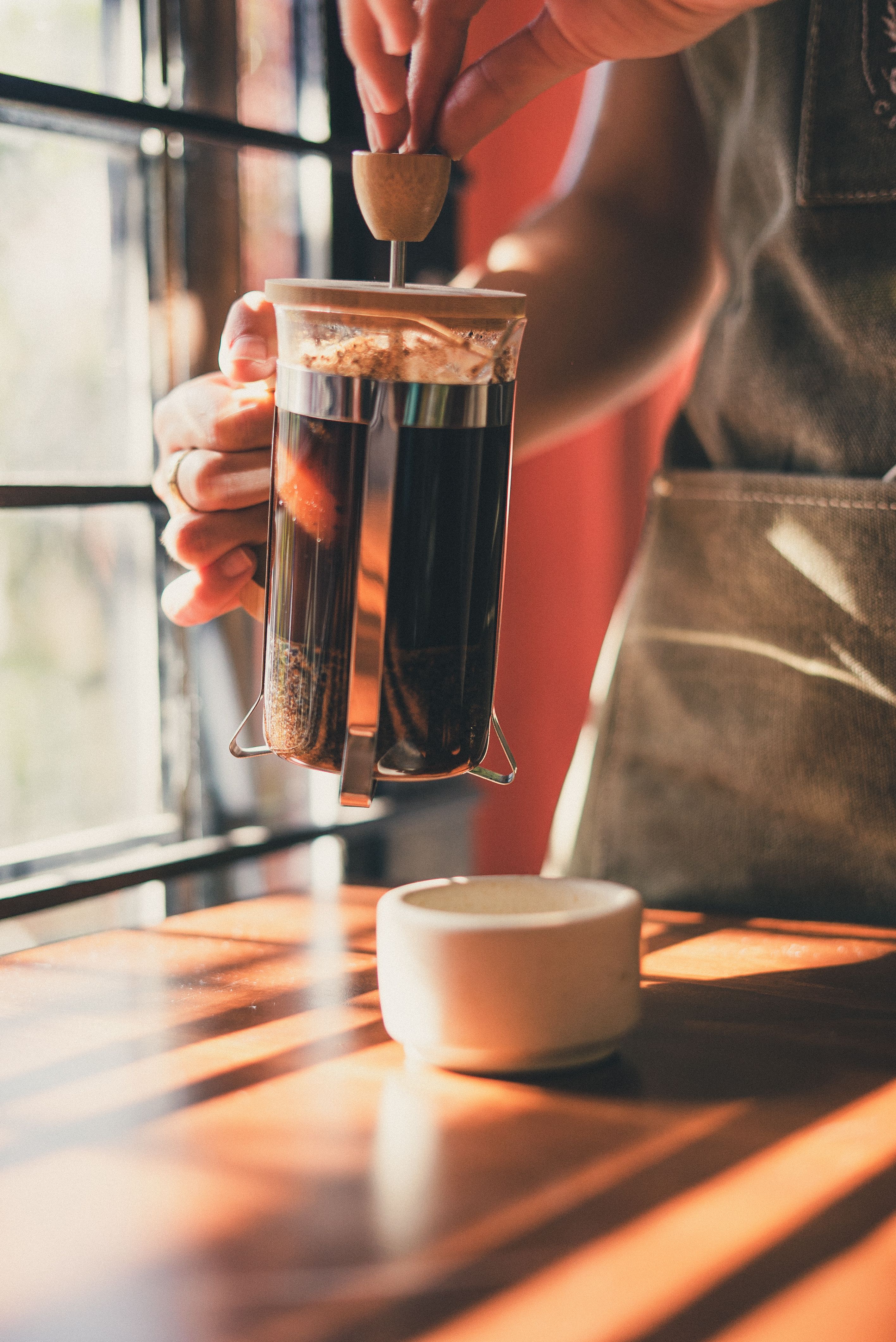 A barista uses a French press to brew coffee in a well-lit cafe.